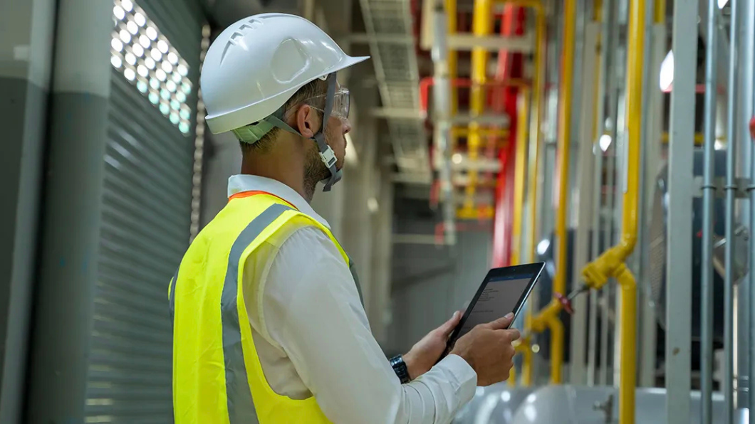 A person wearing a hardhat and safety vest inside an indoor area with industrial equipment and pipes inspects the area and makes notes on a tablet.