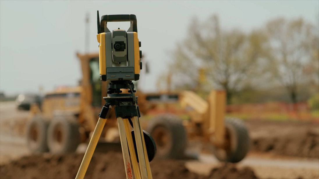 A surveying instrument on a tripod in front of a grader on a construction site.
