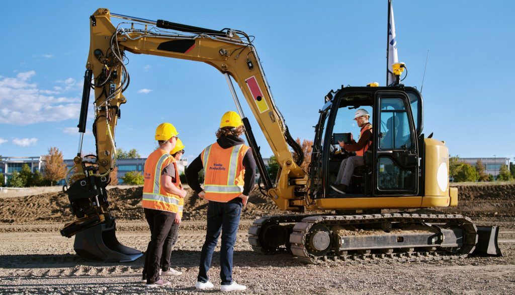 Construction workers in hard hats and safety vests stand near an excavator on a sunny day.