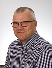 Markku Raisanen, wearing glasses and a checkered shirt, smiles at the camera.