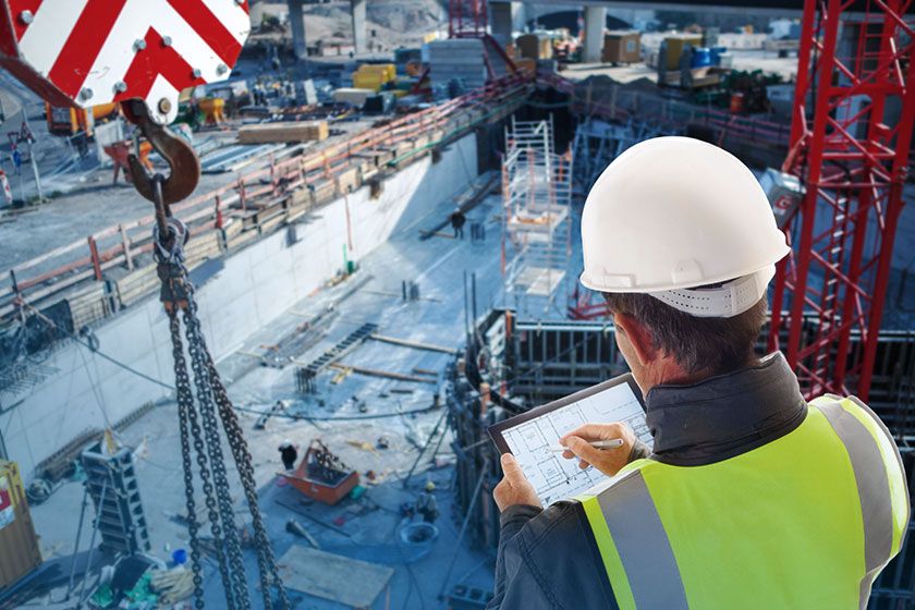 A construction worker in a hard hat and safety vest reviews blueprints on a tablet at a busy construction site with cranes and heavy machinery.