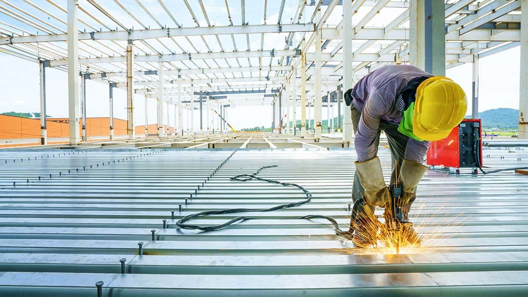 Worker in yellow hard hat welding on metal decking, creating sparks inside an open steel-frame building.