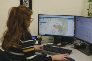 Woman with auburn hair wearing glasses looks at two computer screens displaying maps and data.