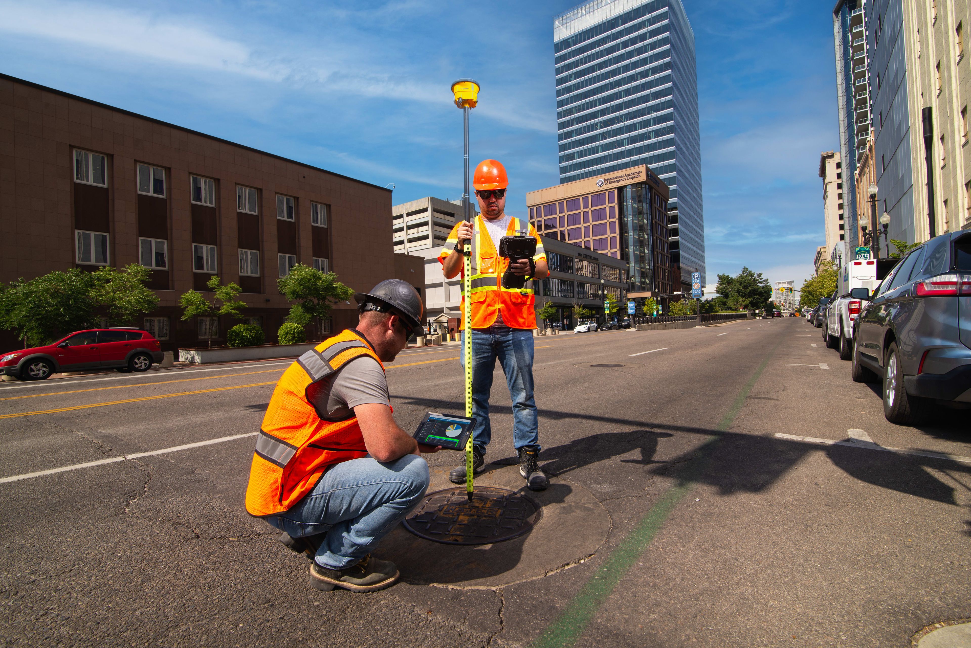 Two surveyors in hard hats and high-vis vests use GPS equipment on a city street.