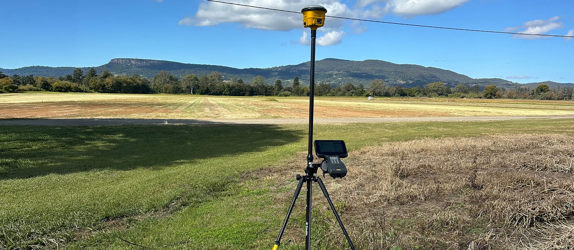 Trimble GNSS receiver mounted on a tripod set up in a field overlooking distant hills.