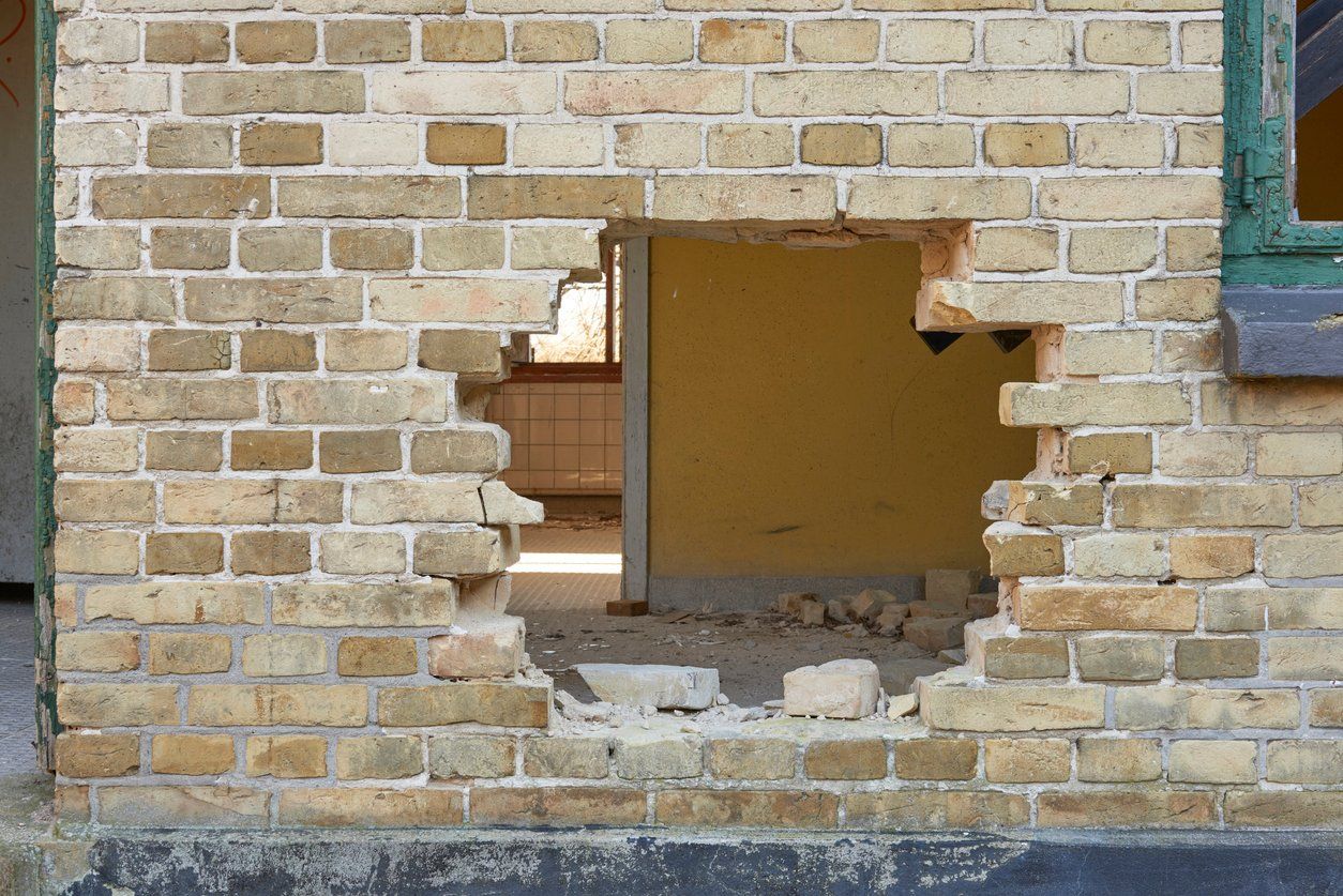 Brick wall with a large hole revealing a yellow room, debris, and a green window frame.
