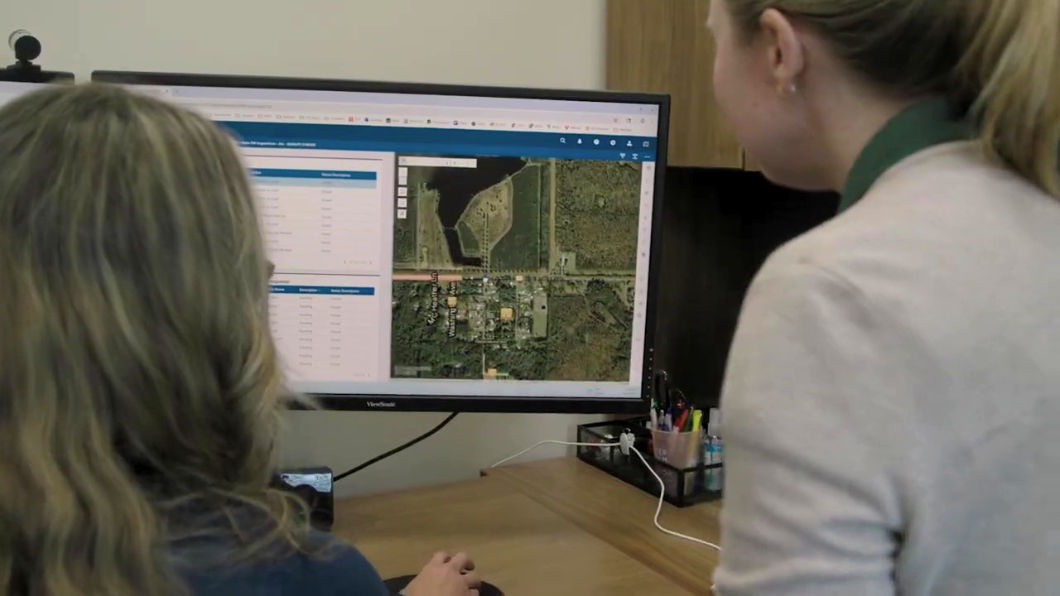Two office workers analyze a digital satellite map on a computer monitor using geographic information system (GIS) software to track county data.