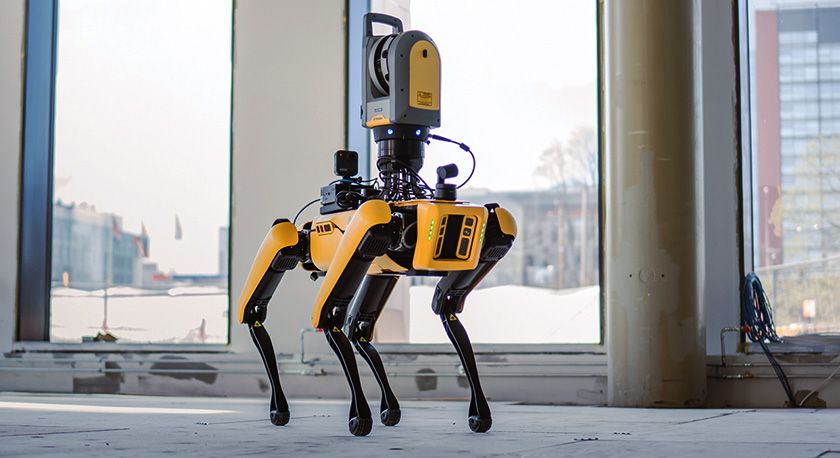 A yellow and black robotic dog with a scanner on its back stands on a concrete floor in a large, unfinished room.