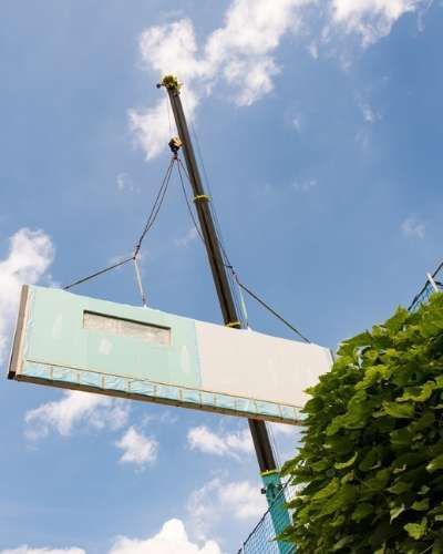 A modular wall panel is lifted into place by a crane against a bright blue sky with fluffy white clouds.