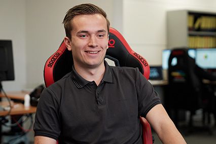 Junger Mann in schwarzem Poloshirt sitzt lächelnd in einem rot-schwarzen Gaming-Stuhl in einem Büroraum.
