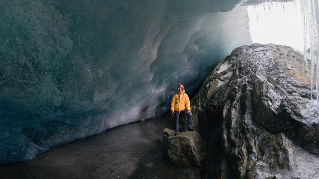 Klaus Thymann in a yellow jacket standing on a rock inside a vast ice cave with icicles at the entrance.
