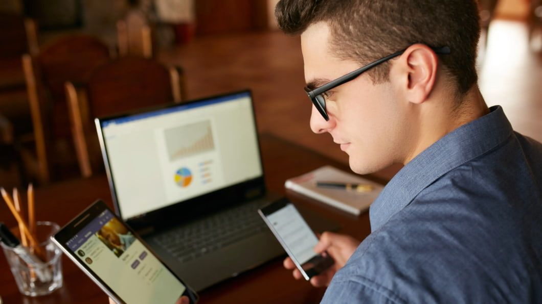 A person with glasses holds a tablet and smartphone while sitting at a wooden desk with an open laptop showing charts.