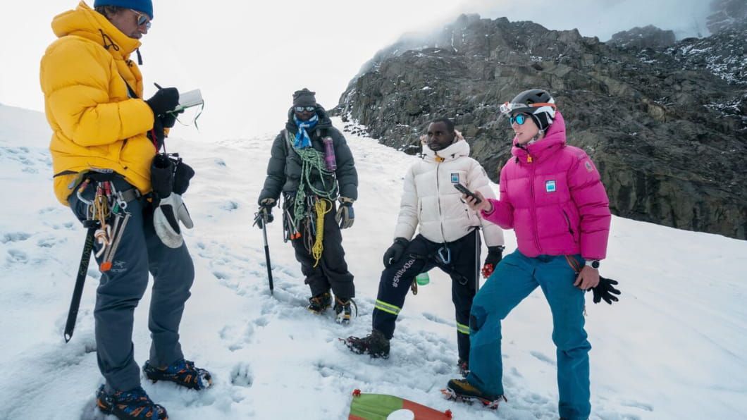 Four mountaineers in colorful gear standing on a snowy slope, equipped with climbing tools and ropes.