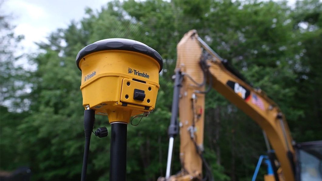 Yellow Trimble GNSS receiver mounted on a pole, with a yellow CAT excavator and green trees blurred in the background.