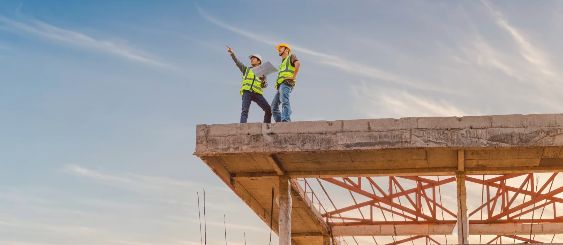 Twee bouwvakkers wijzen vanaf de top van een betonnen constructie.