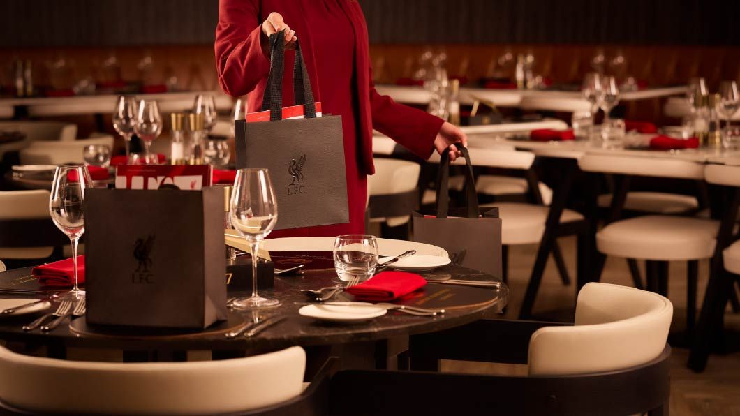 Person in red holding LFC branded gift bags at a formally set dining table with wine glasses and red napkins.