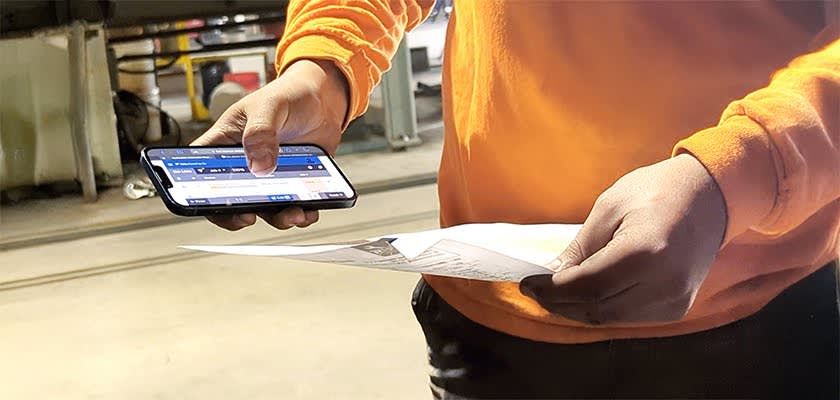 Worker in orange shirt using smartphone and holding paper documents.