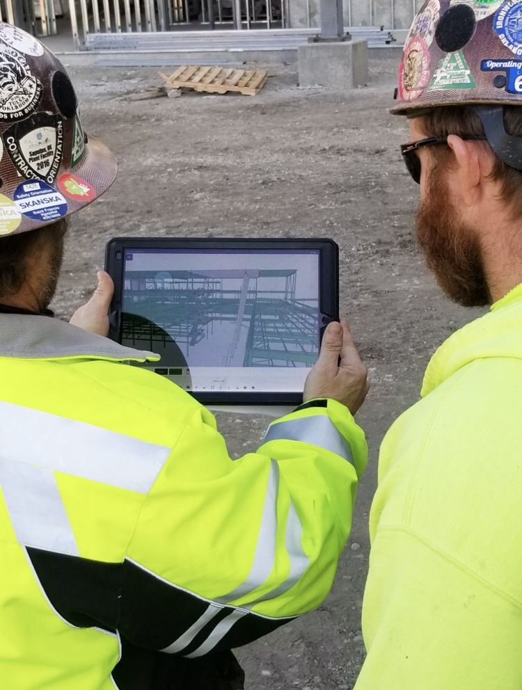 Two construction workers in hard hats and high-visibility vests look at a tablet displaying a 3D model of a building.