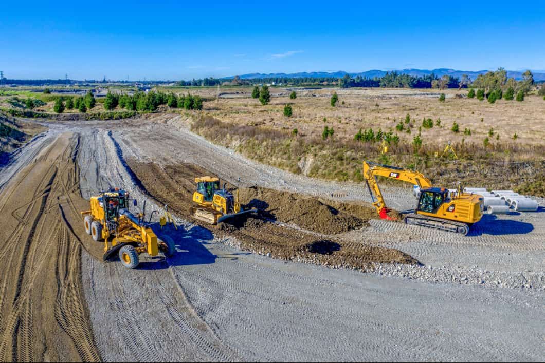 Aerial view of a construction site. Grader, bulldozer, and excavator work on a wide dirt road. Distant mountains under blue sky.