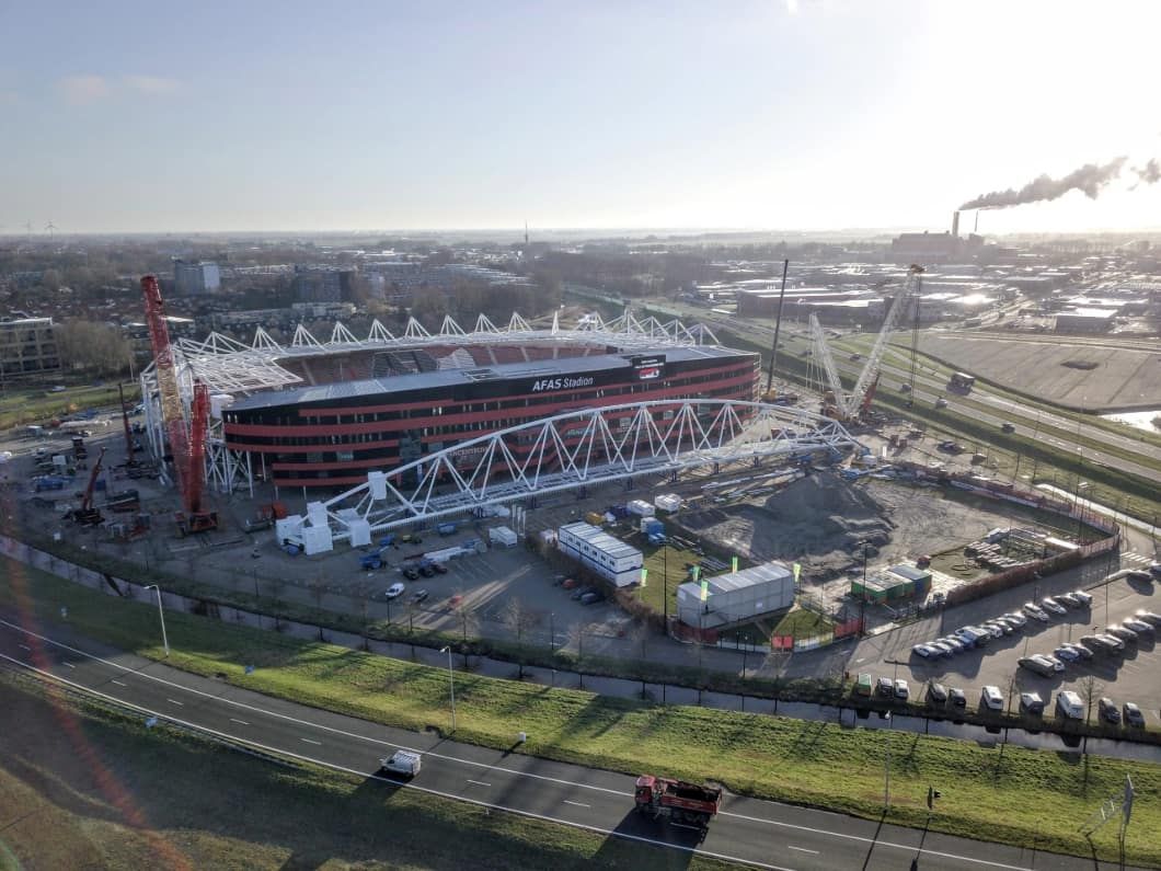 Construction of a modern stadium with red and black exterior, featuring large white truss structures and cranes on a sunny day.