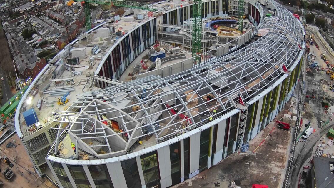 Aerial view of a large, curved building under construction, with exposed metal framework for the roof and visible construction activity.