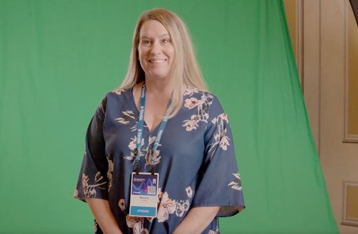 A smiling woman in a blue floral dress and conference badge stands in front of a green screen.