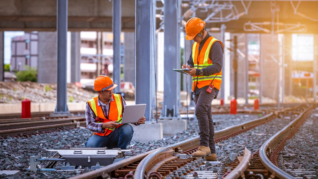 Two people wearing hardhats and safety vests at a newly constructed railway inspect the work and record measurements on their laptop and tablet.