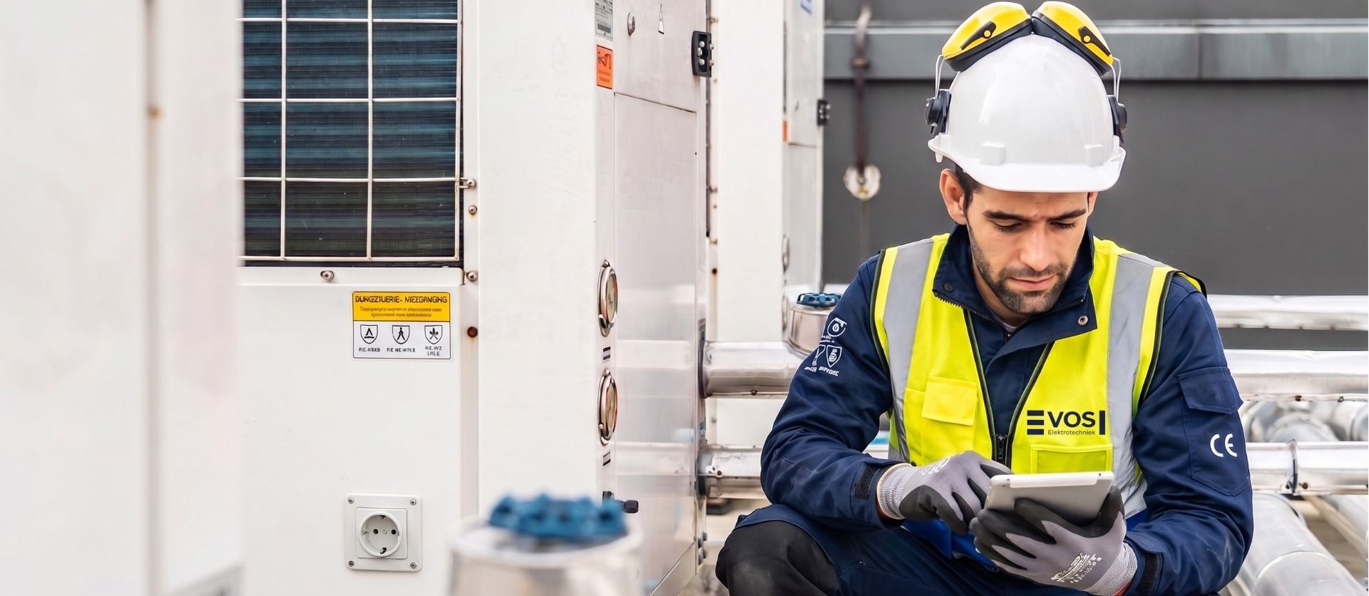 A VOS Elektrotechniek technician in a white hard hat and yellow safety vest uses a tablet near industrial HVAC equipment.