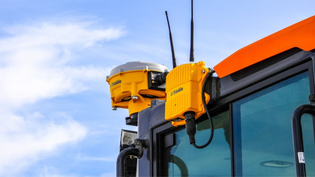 Yellow Trimble GPS receiver and antenna mounted on top of an orange construction machine cab against a blue sky.
