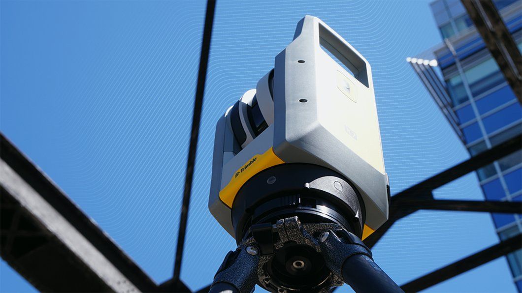 Gray/yellow Trimble X7 3D laser scanner on tripod, surveying a modern building for AECO/construction data.