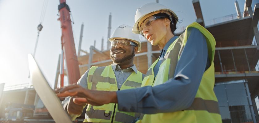 Two construction workers in hard hats and vests look at a laptop on a sunny construction site with a crane.