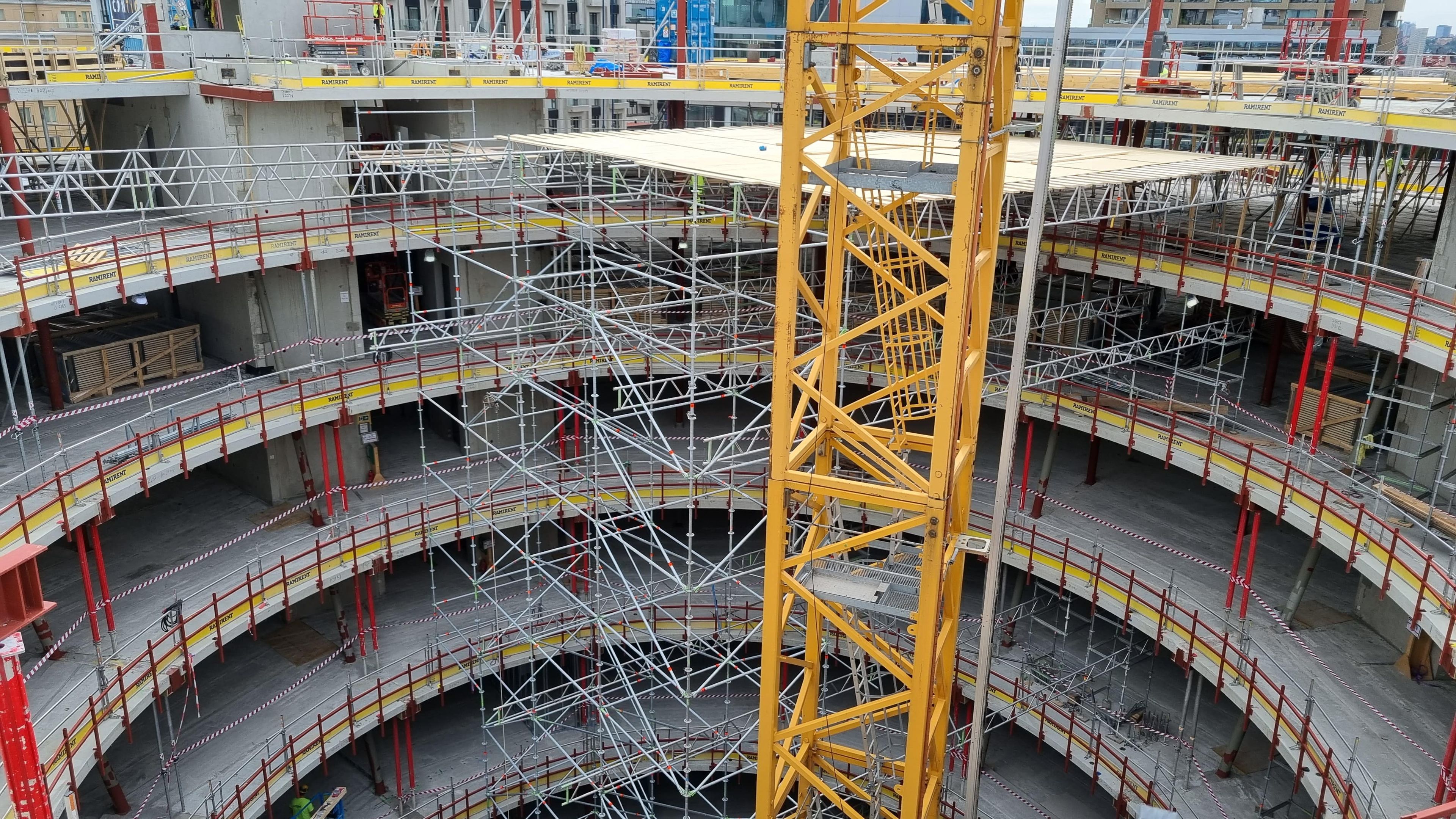 A crane arm is visible against scaffolding and concrete structures of a large, multi-story building under construction.
