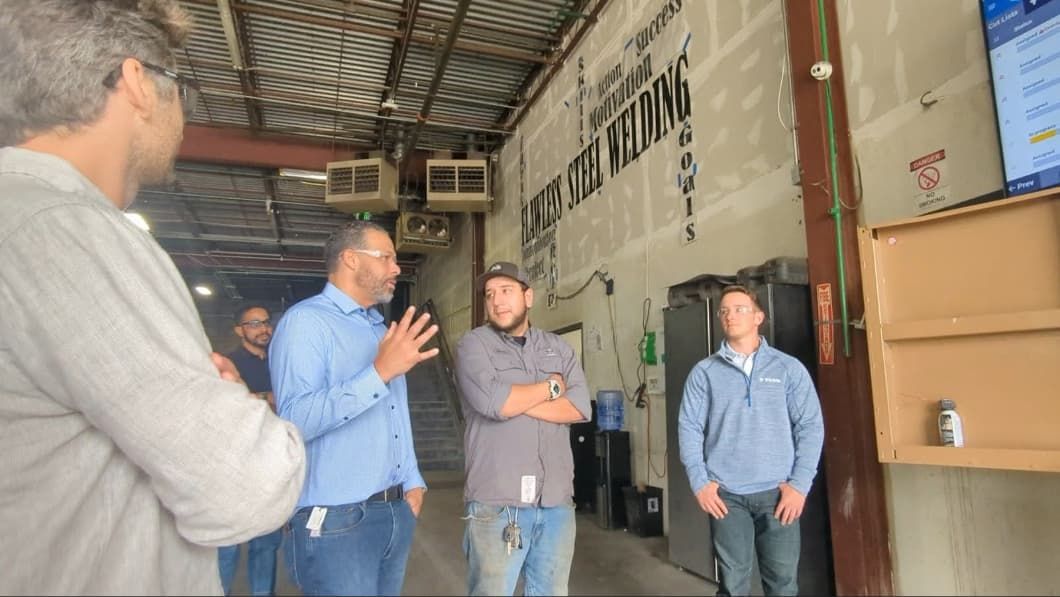 A group of men stand in a workshop, listening to a speaker. Motivational words are painted on the wall behind them.