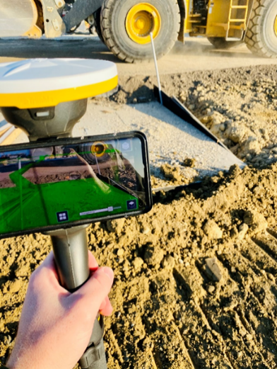 A person holds a smartphone displaying an AR overlay over a construction site.
