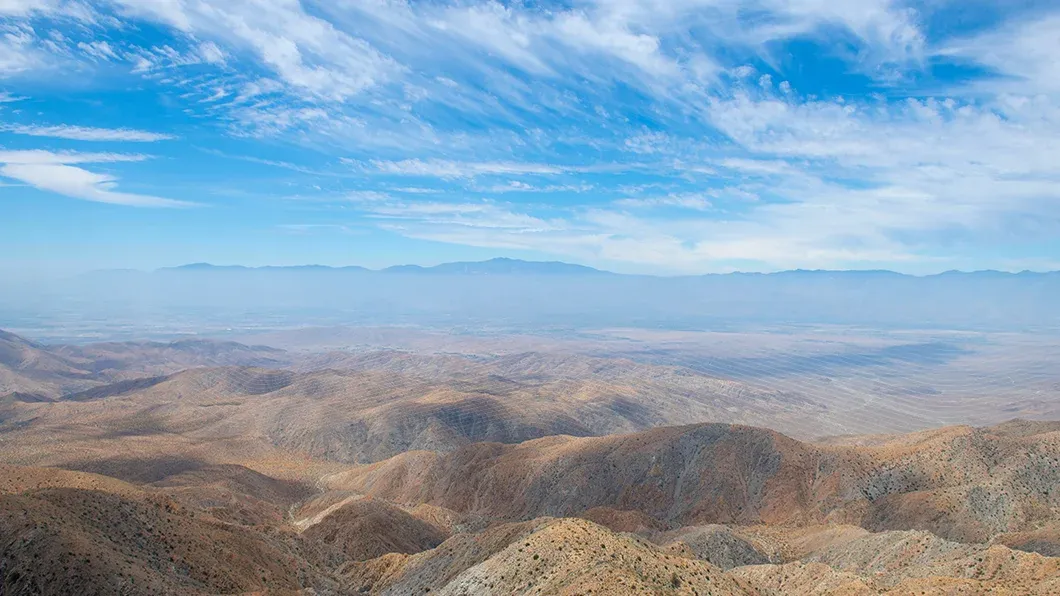 High-angle panoramic view of desert hills and the Coachella Valley from Keys View in Joshua Tree National Park under a blue sky with wispy clouds.