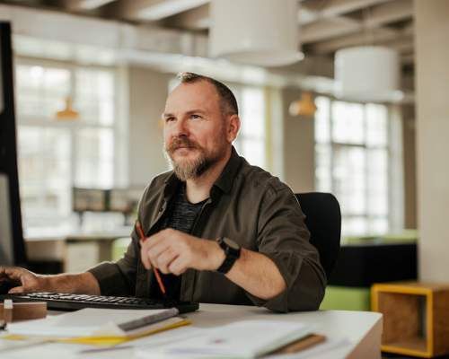 Man sitting in front of computer in an open office setting with lots of windows.
