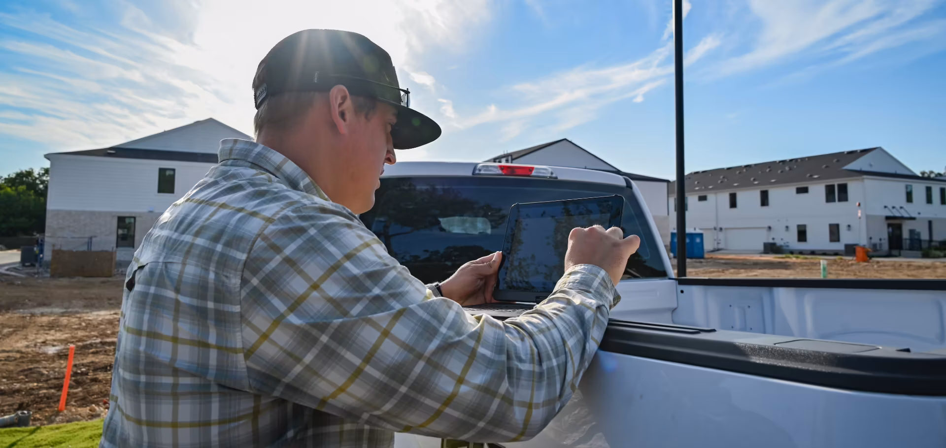 Man in a cap and plaid shirt uses a tablet on the back of a white pickup truck at a construction site with partially built houses.