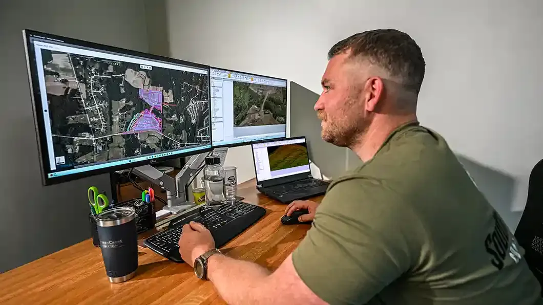 Man sitting at a desk looking at 3D modeling and site mapping software on multiple wide monitors.
