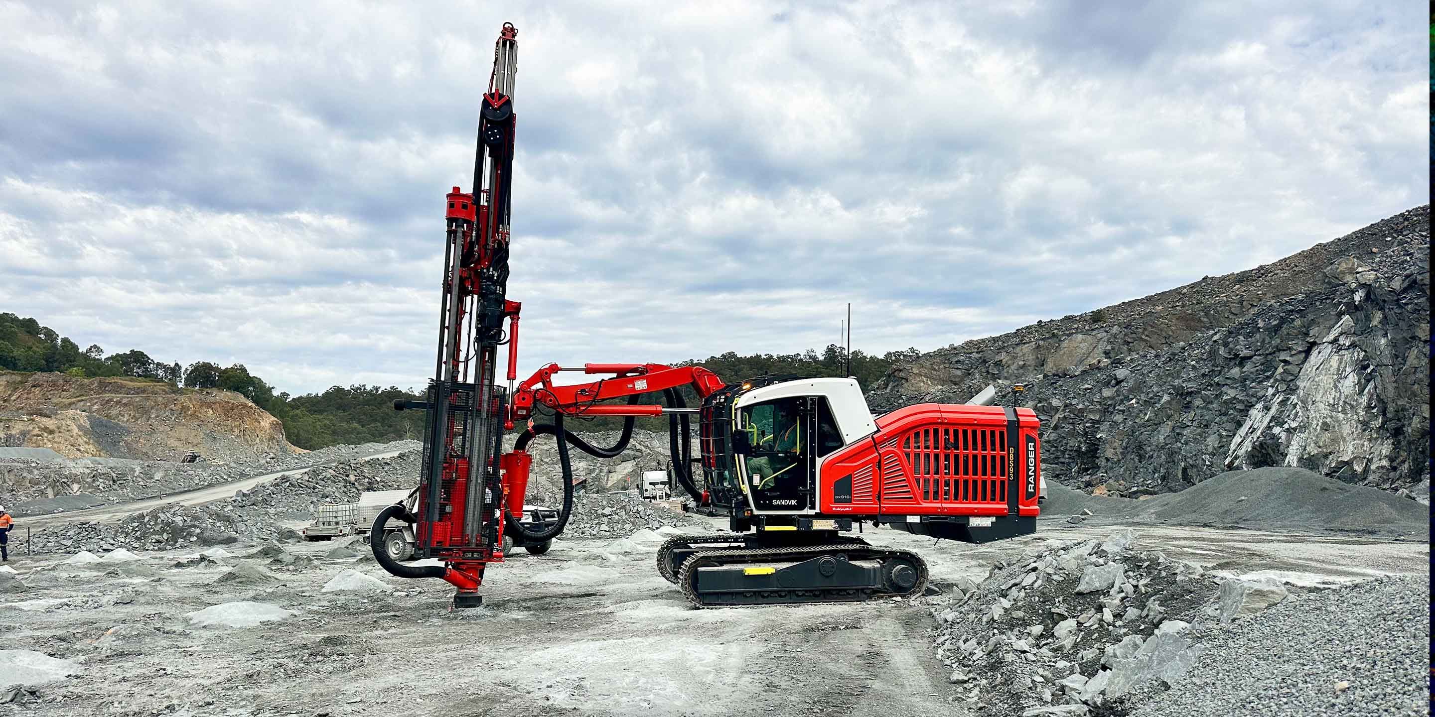 Side view of a red drill rig positioned on cleared rocky terrain in an open-pit quarry.