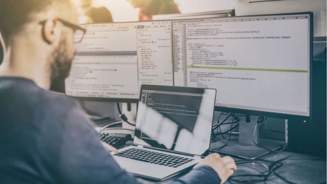 A person works at a desk with a laptop and multiple monitors displaying code, in a modern, well-lit tech workspace.