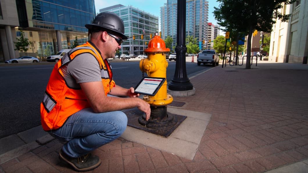 Construction worker in orange safety vest using tablet while crouched next to yellow fire hydrant on city sidewalk.