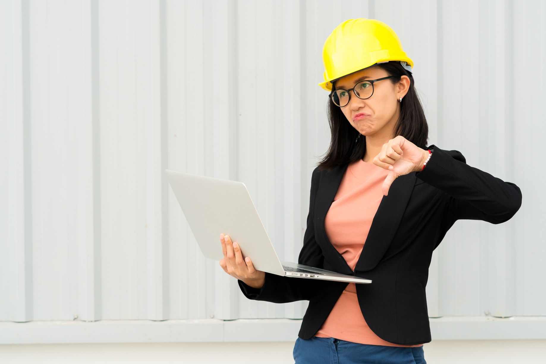 A woman wearing a yellow hard hat and glasses holds a laptop and gives a thumbs down.