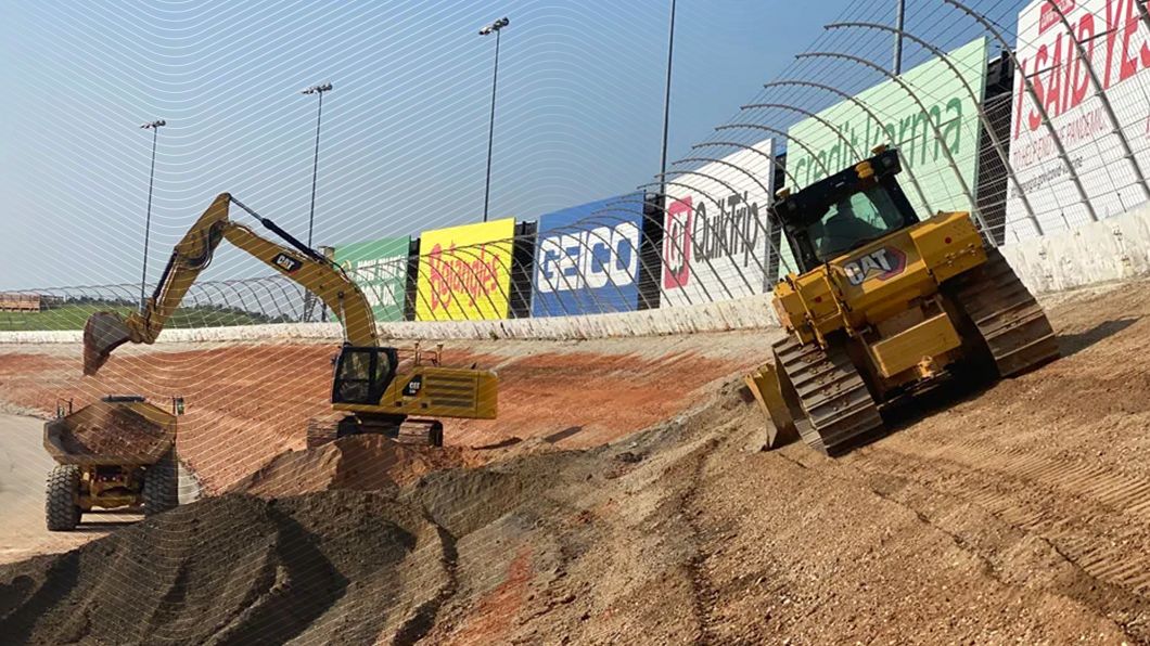 Yellow Cat excavator and bulldozer perform earthmoving on a banked NASCAR track, featuring sponsor billboards for Kroger, GEICO, and Credit Karma.