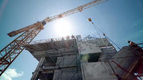 A yellow crane looms over a concrete building under construction, with workers visible on the upper levels.
