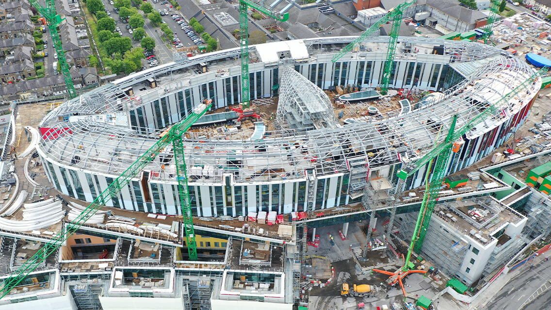 Aerial view of the St. James's Hospital structure under construction in Dublin, with multiple green cranes surrounding the oval-shaped building.