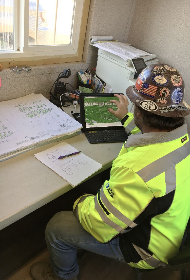 A construction worker in a neon yellow jacket and hard hat studies a digital rendering on a laptop, with large stacks of blueprints nearby.