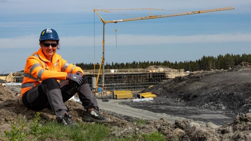 Woman in construction gear sits on a hill at a construction site with a crane.