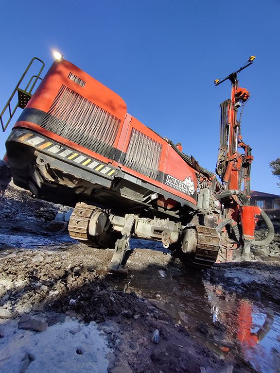 Orange drilling rig with tracks operating in muddy terrain under a clear blue sky.
