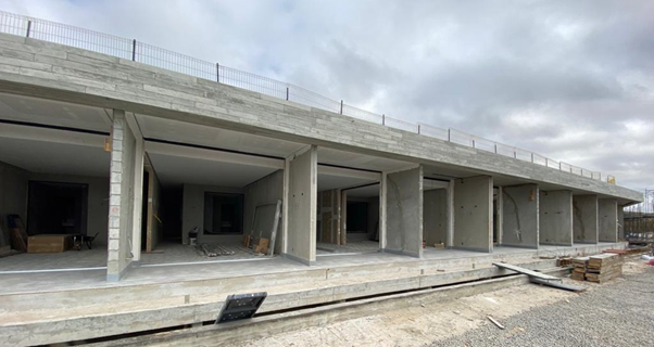 A long, unfinished concrete building with multiple open doorways and a wire fence along the top edge under a cloudy sky.