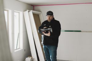 A man in a black jacket and blue jeans holding a black bag in a room under construction.